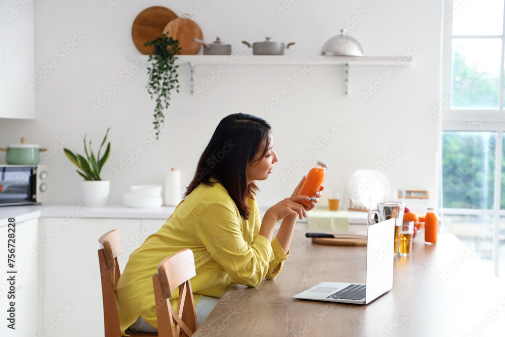 Beautiful Asian woman with bottle of juice and laptop in kitchen Beautiful Asian woman with bottle of juice and laptop in kitchen