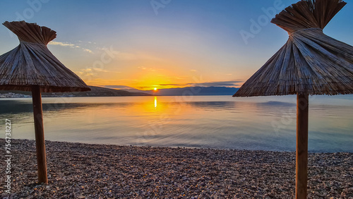 Straw umbrellas at sunset on idyllic pebble beach in tourist resort town Baska, Krk Otok, Primorje-Gorski Kotar, Croatia, Europe. Paradise summer atmosphere in Kvarner bay, Mediterranean Adriatic Sea