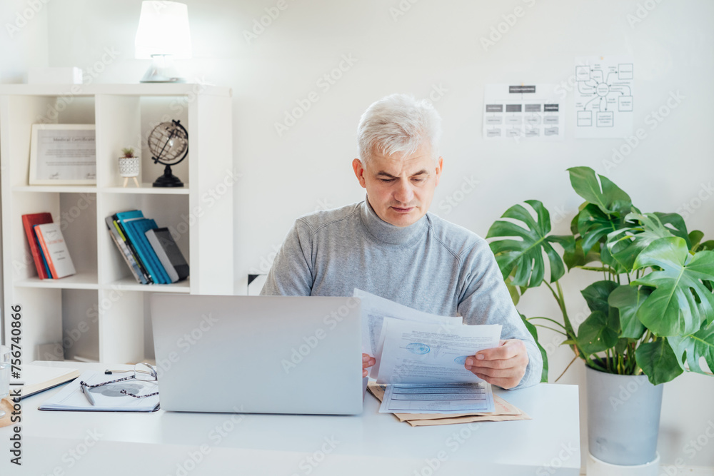 Middle aged businessman reading paper documents and using laptop while working at home office. Confident, experienced senior male professional. Small business entrepreneur manage business paperwork