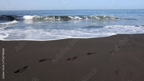 Ocean waves hitting volcanic black sand beach with human footprints. No visible people