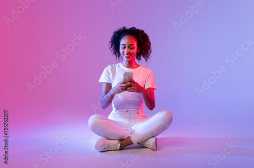 Happy black lady using phone, sitting cross-legged on neon background