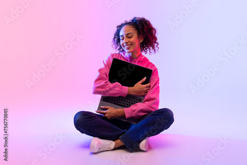 Content black lady hugging laptop with joy on vivid gradient backdrop