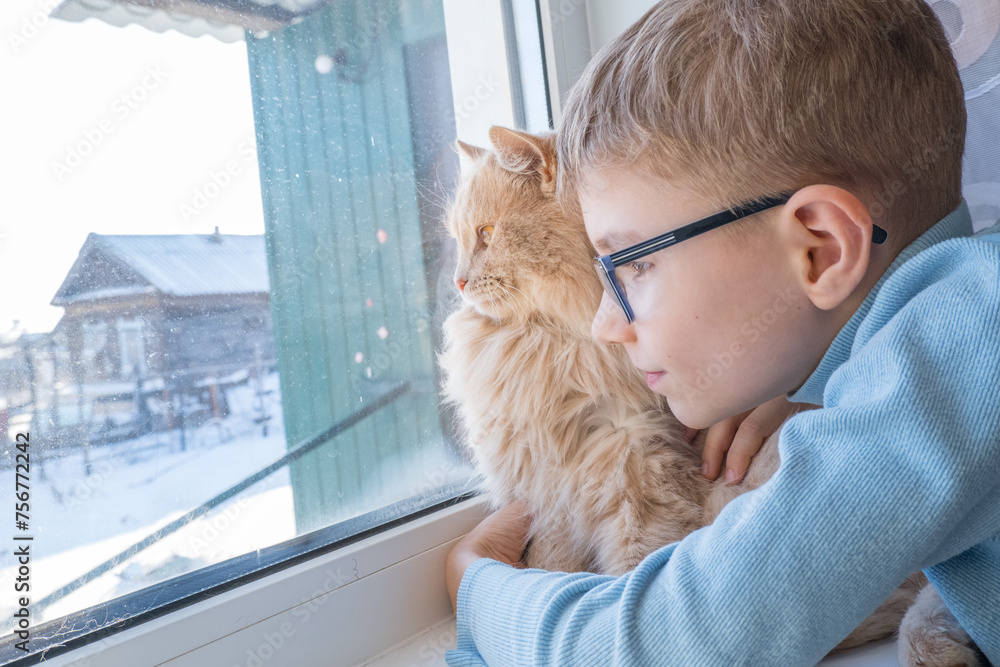 Beautiful scene two friends looking out fo window. Small boy with a cat stares out of window watching falling snow, focus on cat