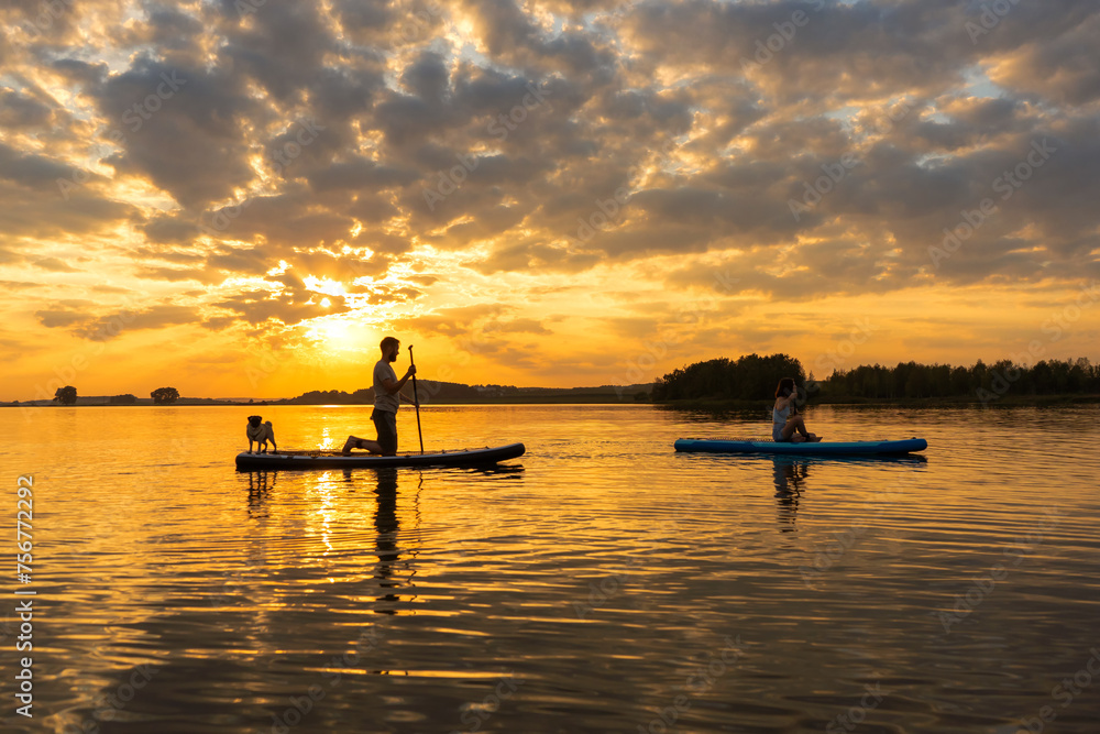 Happy couple paddle boarding at lake during sunset together with pug dog. Concept of active family tourism and supping with pets. Brave Dog Standing on SUP Board and enjoy lifestyle on summer vacation