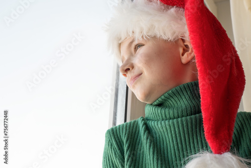 Obraz na płótnie Children's dreams concept. Friendship. A lonely little boy in a Santa Claus hat is sad at the window waiting for gifts, looking out the window, the child is waiting for Santa Claus. The baby is w salonie