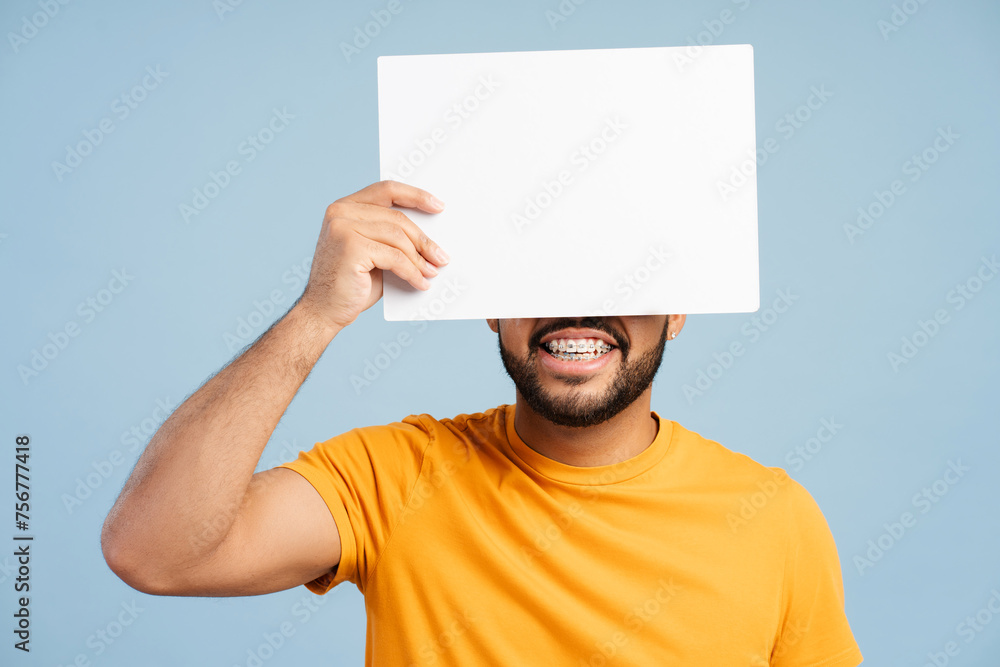 African American man with dental braces, holding a white blank poster and covering his face with it African American man with dental braces, holding a white blank poster and covering his face with it