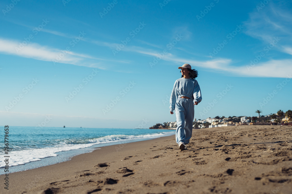 Young woman tourist in blue sport suit walks along the seashore, enjoys the seascape. Travel, tourism concept. Active lifestyle.