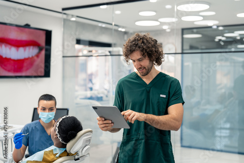 Handsome male dentist in green uniform working on digital tablet in modern dental office with patient in dentist s chair and nurse in background.