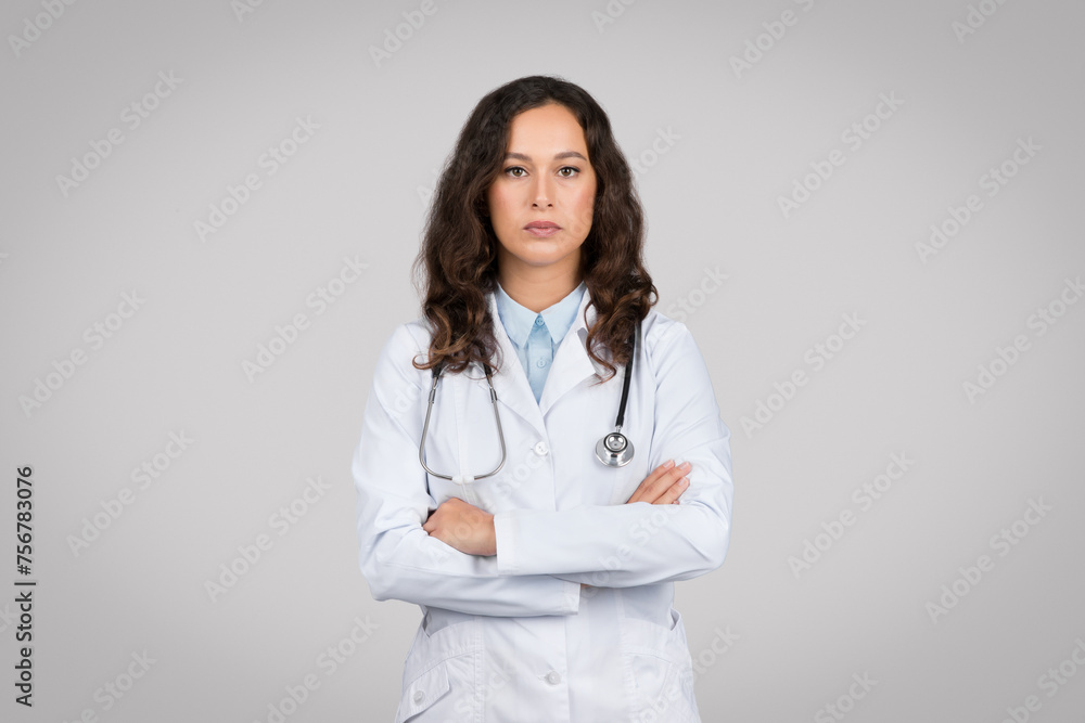 Portrait of confident woman doctor with folded arms, looking at camera, standing against grey background Portrait of confident woman doctor with folded arms, looking at camera, standing against grey background