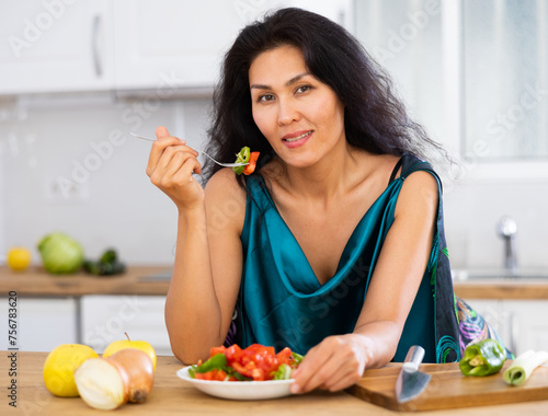 Portrait of cheerful asian woman leaning on table in kitchen and eating just cooked salad. Woman having breakfast with fresh salad.