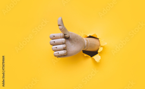 A wooden hand protrudes from a torn hole in yellow paper and shows a big thumbs up like gesture. The concept of approval and that everything will be fine. Artificial intelligence, robot.
