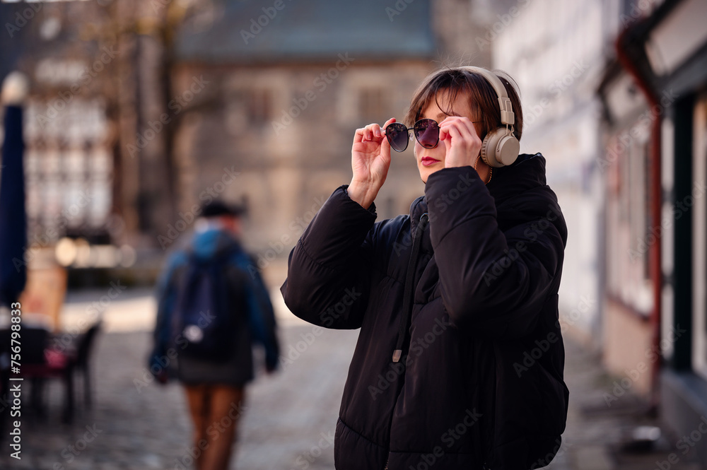In a cobbled street, an elegant woman wearing sunglasses lifts her headphones. Her jacket and expressive stance suggest a blend of modern tech and timeless style In a cobbled street, an elegant woman wearing sunglasses lifts her headphones. Her jacket and expressive stance suggest a blend of modern tech and timeless style