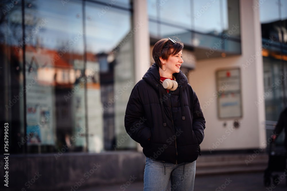 An urban woman stands confidently in front of a reflective glass building, headphones draped around her neck, blending city life and personal leisure