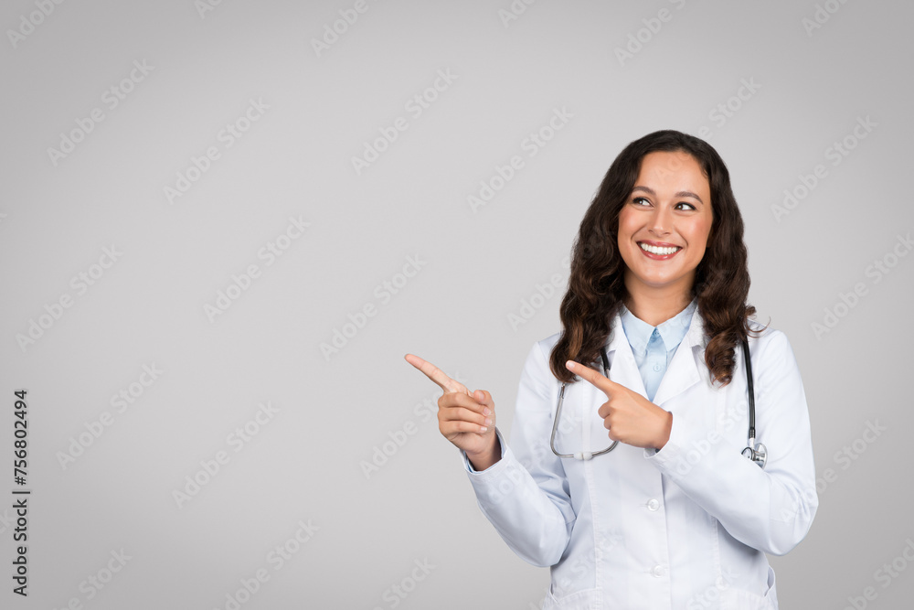 Happy young lady doctor in white coat pointing fingers up at free space, recommending medical ad and offer Happy young lady doctor in white coat pointing fingers up at free space, recommending medical ad and offer