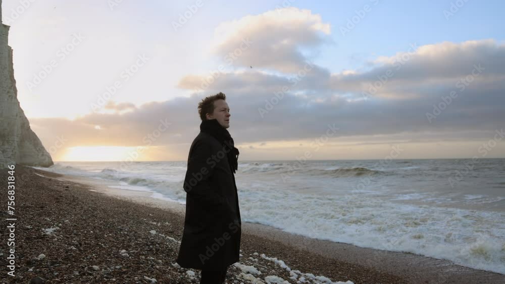 A lone man in a black coat stands on a pebble beach, watching a serene sunrise beside towering chalk cliffs. The scene captures a tranquil morning atmosphere. Seven Sisters, UK, Slow Motion, 4K RAW. A lone man in a black coat stands on a pebble beach, watching a serene sunrise beside towering chalk cliffs. The scene captures a tranquil morning atmosphere. Seven Sisters, UK, Slow Motion, 4K RAW.