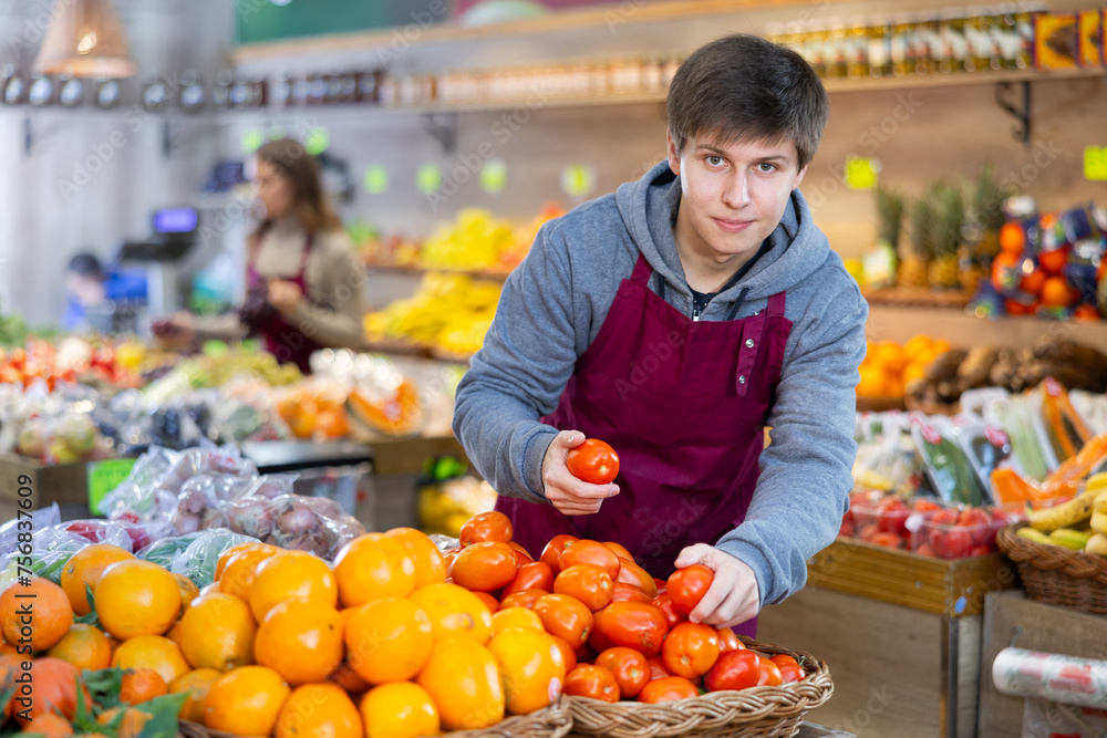 Smiling young salesman putting tomatoes in vegetable basket in large grocery store