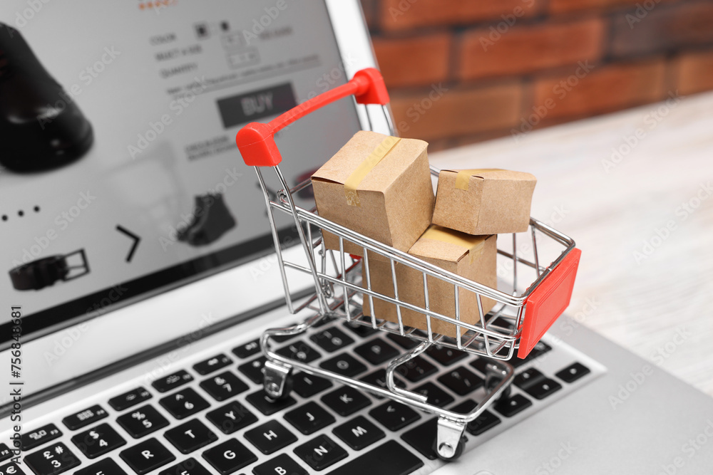 Internet store. Small cardboard boxes, shopping cart and laptop on light wooden table, closeup