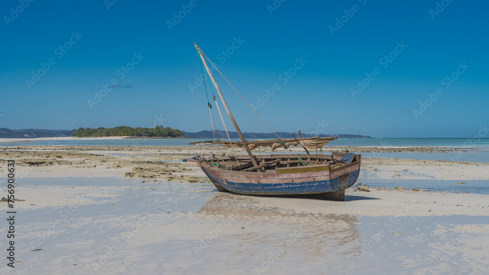 Obraz premium An old weathered fishing boat tilted into shallow water at low tide. A mast, a folded sail against the blue sky. Reflection in a puddle of water. The ocean, a green island in the distance. Madagascar.
