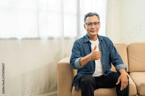 Handsome asian mature old man sitting on sofa couch in living room at home. Happy Portrait of cheerful smiling senior asian man relax in the house. Mature People and lifestyle © Chanakon
