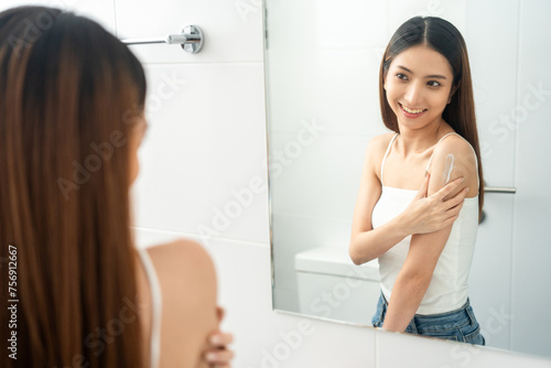 Beautiful smiling happy young asian woman with beauty skin care treatment lotion on the body. Charming female lady looking mirror applying body cream Facial treatment beauty and spa