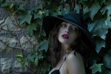 A mysterious woman with a wide-brimmed hat, standing near an old, ivy-covered stone wall. Her eyes hold secrets