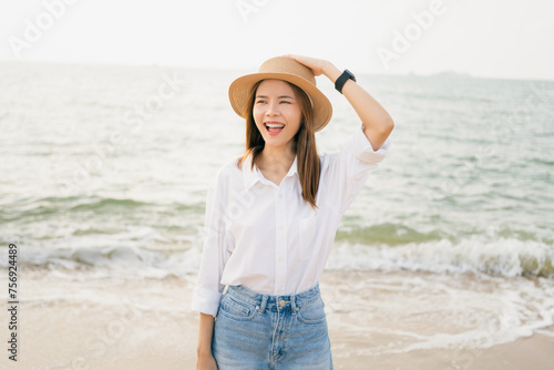 Smiling Asian woman standing on the beach and hand touch hat on head with breaths a full breast and enjoys freedom. Summer time concept.