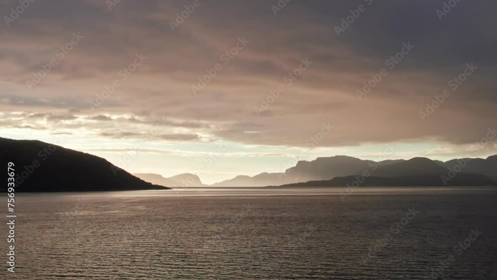 Panoramic aerial shot, sweeping over Sognefjord in Norway towards a bright sunset over the mountainous horizon. The sky is dark and cloudy. Panoramic aerial shot, sweeping over Sognefjord in Norway towards a bright sunset over the mountainous horizon. The sky is dark and cloudy.