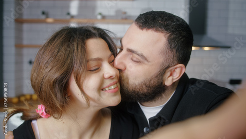 Young couple in love making a selfie using phone having fun in the kitchen looking at camera smiling. Man kiss to girl