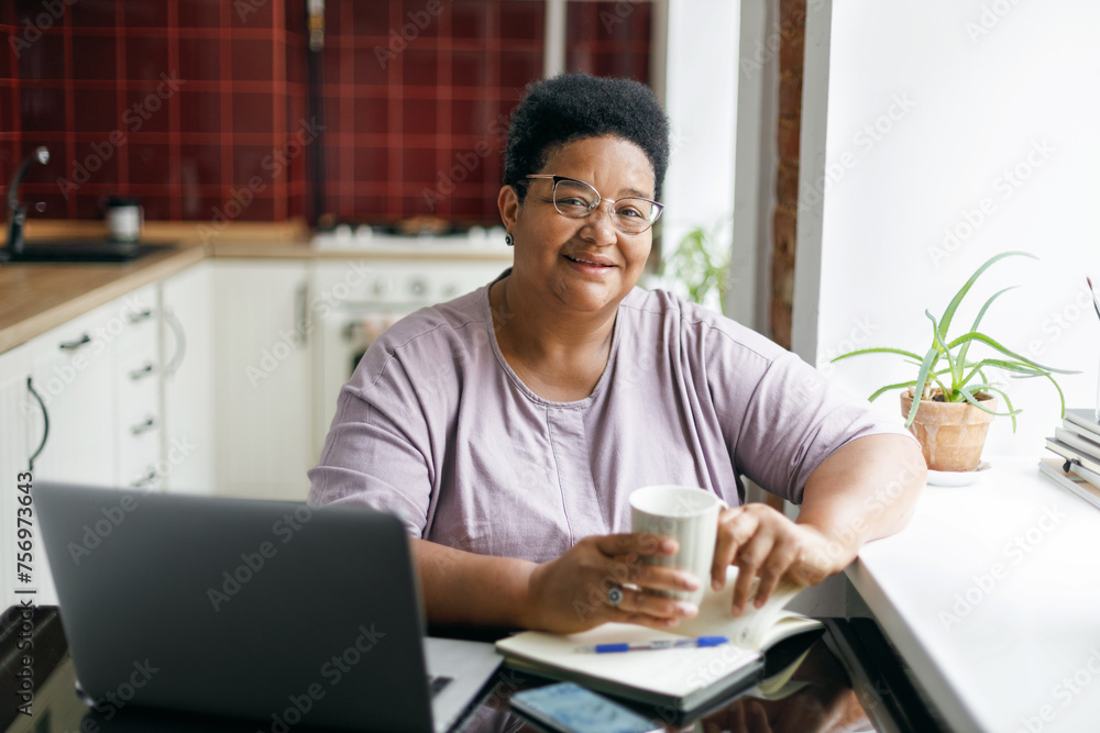 Happy cheerful retired african american plus size female in glasses holding cup of coffee sitting at kitchen near window in front of laptop, browsing internet and websites with culinary recipes Happy cheerful retired african american plus size female in glasses holding cup of coffee sitting at kitchen near window in front of laptop, browsing internet and websites with culinary recipes