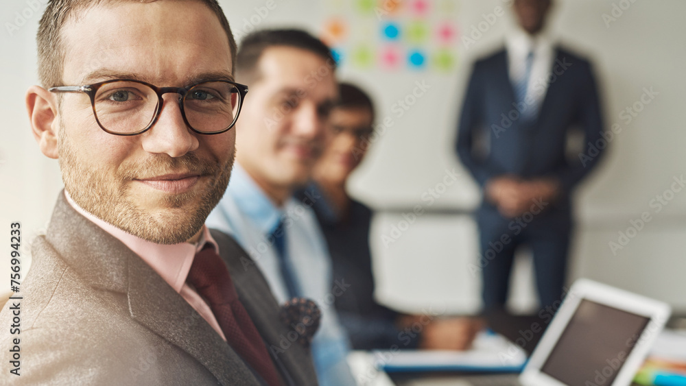 Close up portrait of a smiling businessman or CEO with glasses looking into the camera. His team of diverse colleagues can be seen in the background Close up portrait of a smiling businessman or CEO with glasses looking into the camera. His team of diverse colleagues can be seen in the background
