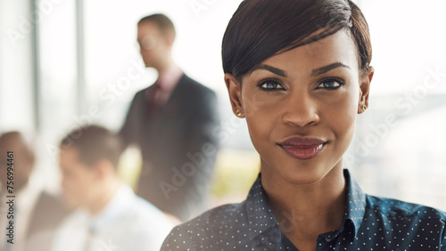 Close up portrait of a black confident business woman looking directly into the camera. Standing in a meeting room with colleagues behind her photo