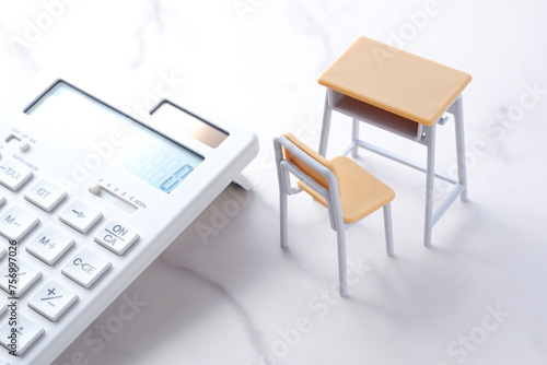 学校の机と椅子、電卓がイメージする、高騰を続けて家計を圧迫する教育費のイメージ 