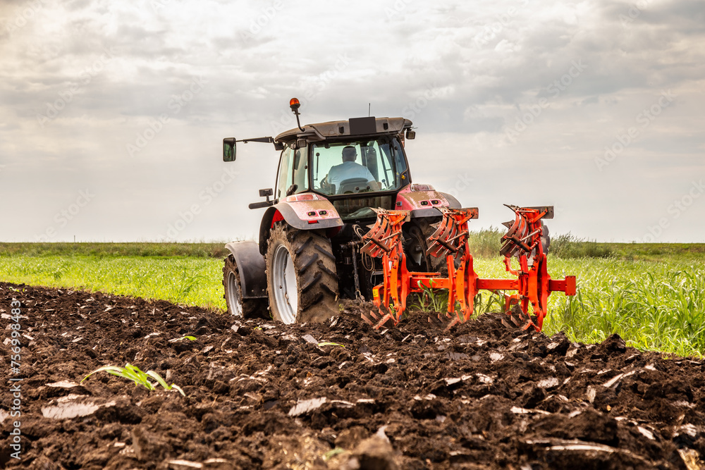 Red tractor is engaged in plowing the soil in a vast green farmland under a cloudy sky Red tractor is engaged in plowing the soil in a vast green farmland under a cloudy sky