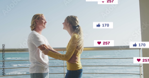 Image of social media icons on banners over caucasian couple in love embracing by seaside