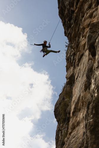 A man is jumping off a cliff with a rope. The sky is blue and the clouds are white