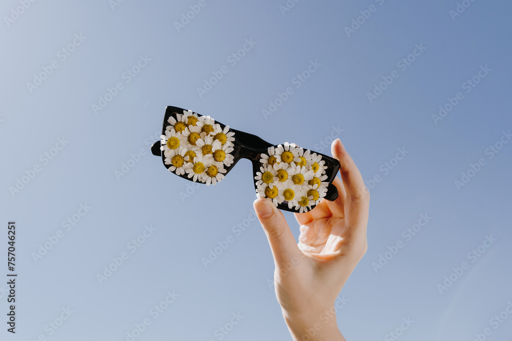 Hand holding sunglasses decorated with chamomile daisy flower buds over blue sky. Spring, summer sunny day concept with copy space for text - obrazy, fototapety, plakaty 