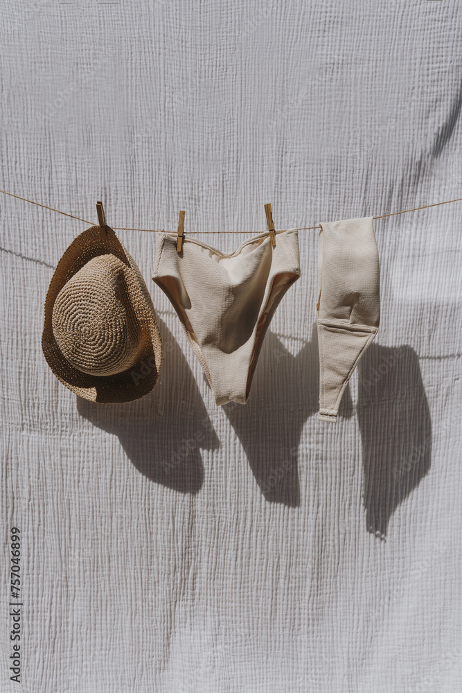 Female swimwear and straw hat hanging over white cotton cloth with strong shadows. Sunbathing on a summer sunny day concept - obrazy, fototapety, plakaty 