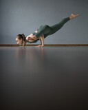 Young woman doing yoga in gym on mat. Koundinyasana. Sage Kaundinya's pose. Flying Splits. Holding your body on your palms. Yoga-coach. balancing pose. Gray background. Reflective floor. Side view. 