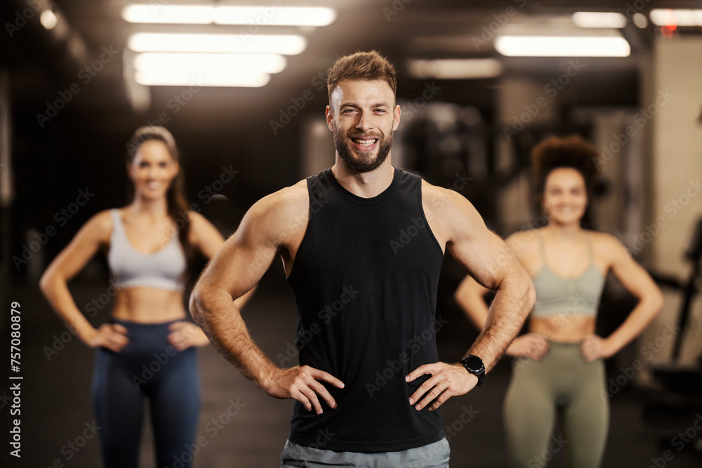 A strong sportsman is confidently posing in a gym and smiling at the camera. There are sportswomen in a blurry background. A strong sportsman is confidently posing in a gym and smiling at the camera. There are sportswomen in a blurry background.