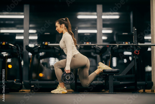 Side view of a strong woman exercising split squats with weight plates in a gym.