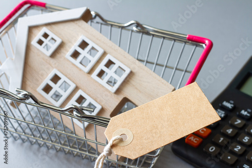 Miniature model of wooden house in shopping basket with tag and calculator, concept of selling, buying or renting real estate