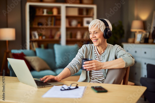 A happy senior adult woman working from home using laptop while enjoying a cup of coffee and listening to music