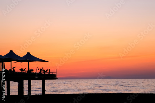 Tropical ocean beach with cafe on beach at dark sunset with purple orange sky. Calm water blue surface and dark shoreline, coast. Travel, tourism, journey, destination on resort for relax concept.