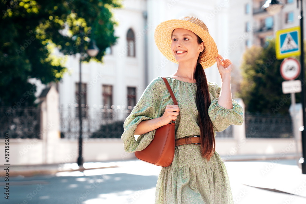 Naklejka premium Young beautiful smiling brunette woman in trendy summer dress. Sexy carefree woman posing in street. Positive model outdoors at sunny day. Cheerful and happy. In hat. Holds handbag