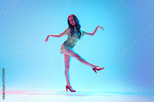 Artistic, talented, beautiful young girl in stylish dress dancing against blue background in neon light. Concept of hobby, dance class, party, 50s, 60s culture, youth photo