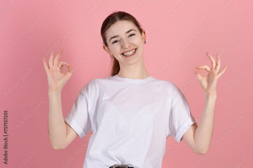Attractive caucasian young brunette woman in casual white t-shirt looks at the camera, smiles and shows okay approve gesture isolated on pink studio background. Attractive caucasian young brunette woman in casual white t-shirt looks at the camera, smiles and shows okay approve gesture isolated on pink studio background.
