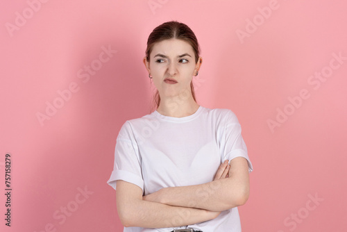 Portrait of a sad brunette girl, Caucasian young woman in a casual white t-shirt with arms crossed, sulking and frowning with disappointed isolated on a pink studio background.