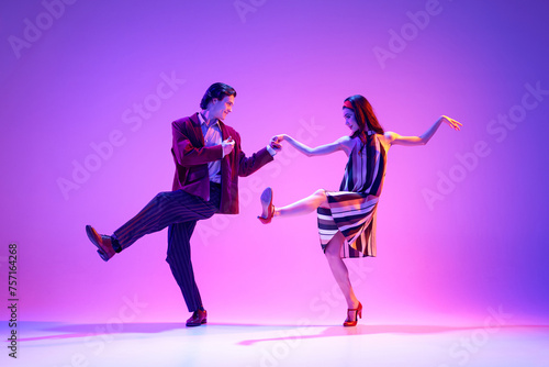 Young man and woman in stylish clothes dancing retro dance, boogie woogie against purple background in neon light. Concept of hobby, dance class, party, 50s, 60s culture, youth
