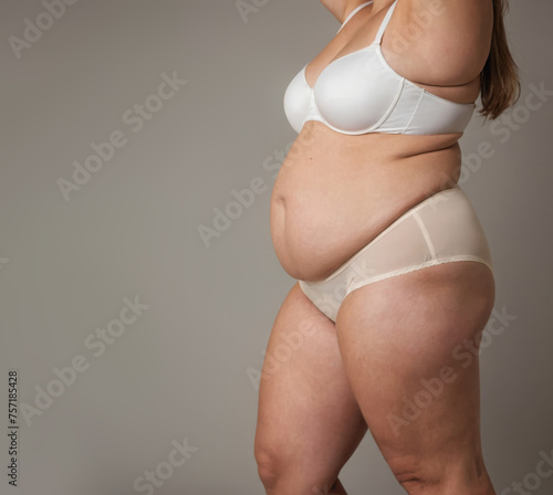 A woman with a white bra and white panties is standing in front of a gray background. Concept of confidence and self-assurance, as the woman is proud of her body and is not afraid to show it