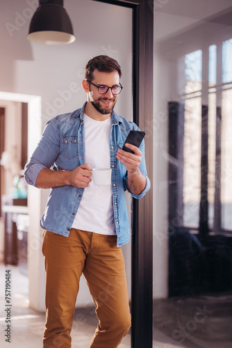 Confident smiling young businessman drinking coffee and using phone while standing in office.